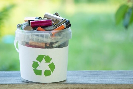 A plastic bin full of batteries. The bin has a green recycling symbol on the label. Greenery is present in the background.