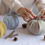 Closeup of hands manipulating cloth into beautiful pumpkin shapes using the Furoshiki method of folding. Pine cones and cinnamon lay on the table as decoration.