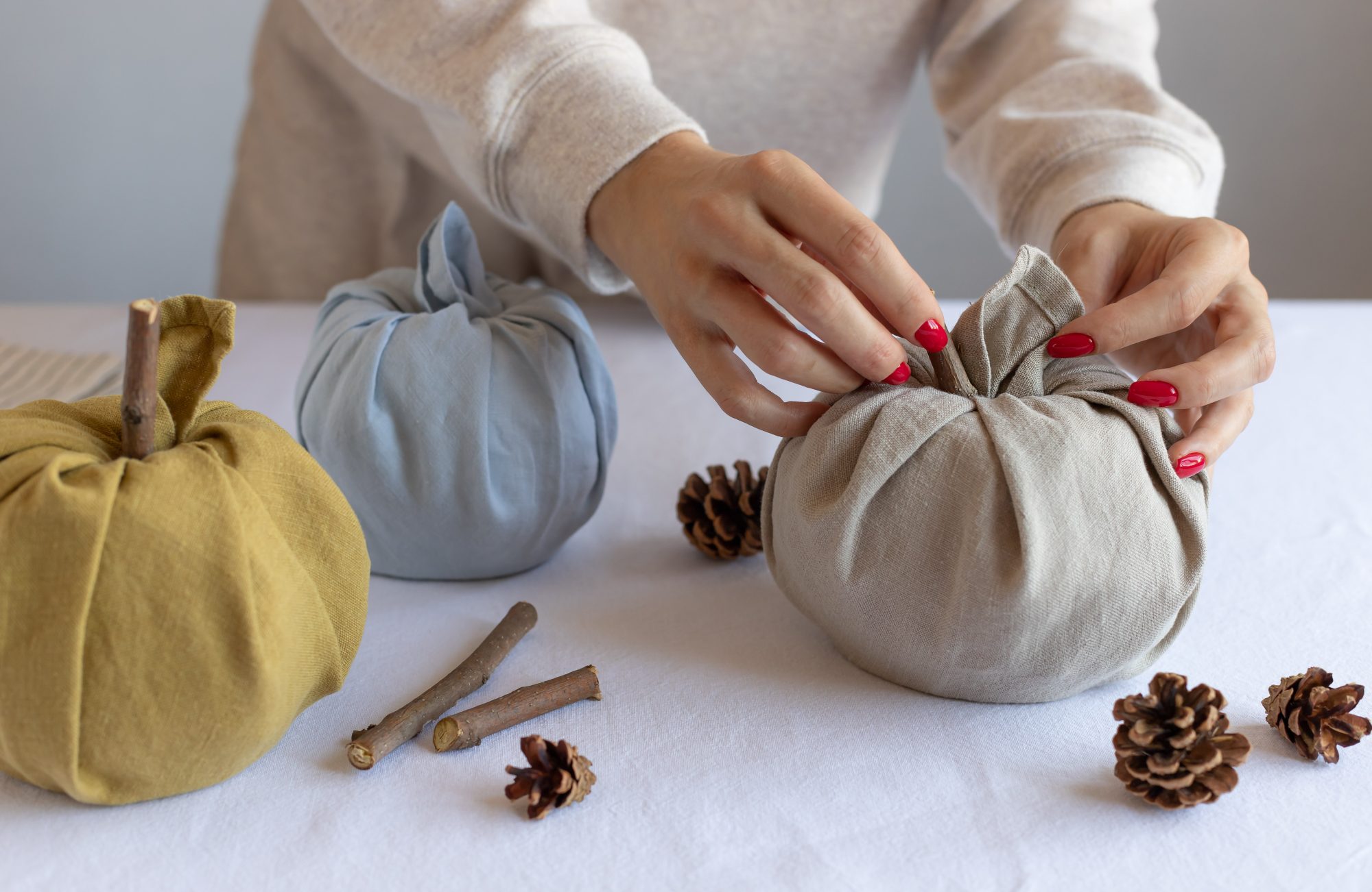 Closeup of hands manipulating cloth into beautiful pumpkin shapes using the Furoshiki method of folding. Pine cones and cinnamon lay on the table as decoration.